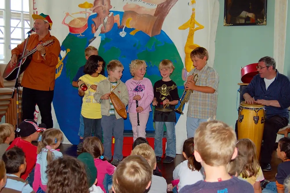 Kinder mir verschiedenen Instrumenten stehen auf einer Bühne, im Vordergrung Kinder, die zuschauen